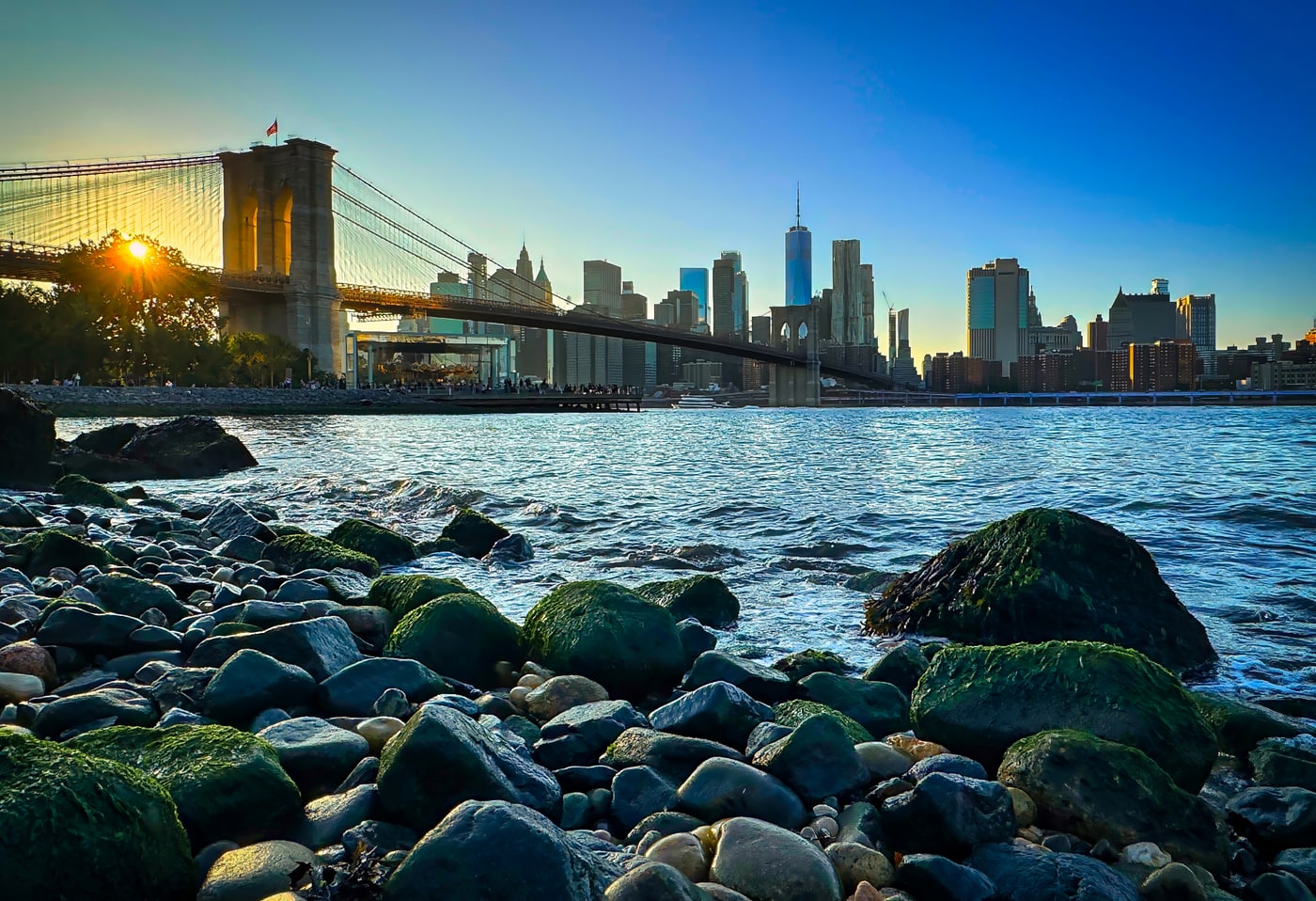 Rocky waterfront with a suspension bridge and skyline in low sunlight.